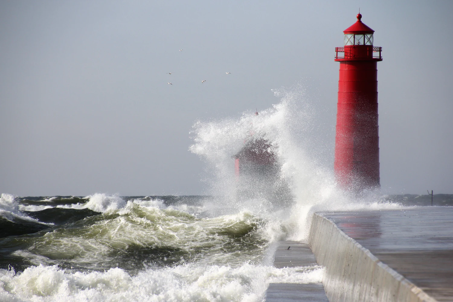 A wave breaking over a pier beside a red lighthouse