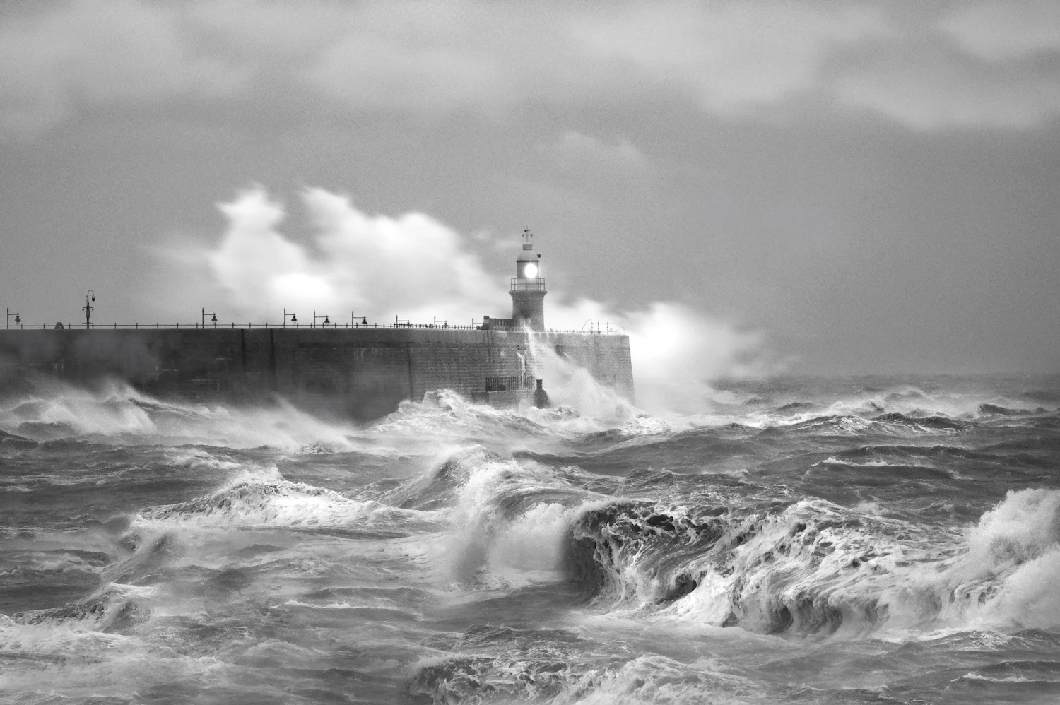 Black and white waves exploding against a pier wall and lighthouse